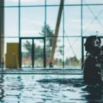 Silhouette of a water polo player mid-action with water splashes inside an indoor pool.