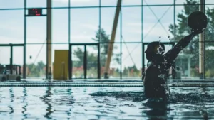 Silhouette of a water polo player mid-action with water splashes inside an indoor pool.