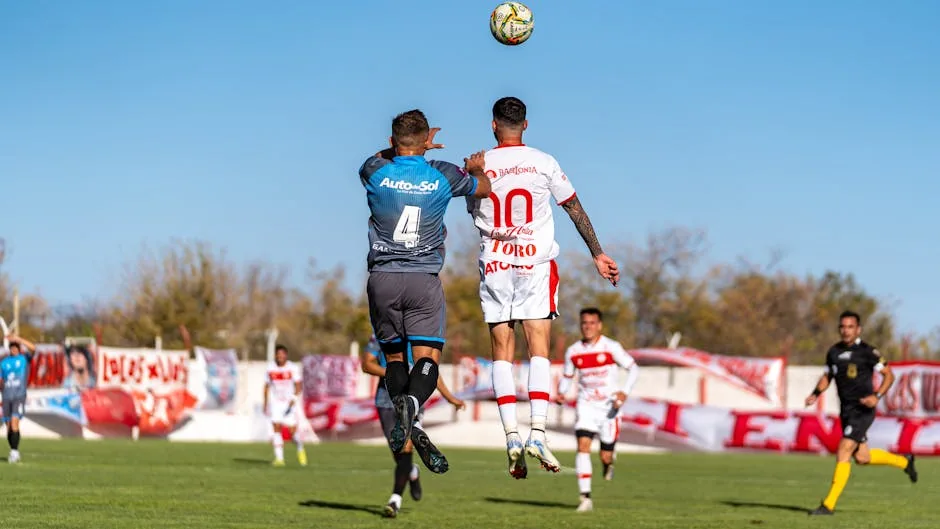 Soccer players in uniforms jumping to head the ball during an outdoor match on a sunny day.